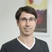 Porträt eines Mannes mit Brille, grauer V-Neck-Sweater, heller Hintergrund und einem farbigen Bild im Rahmen dahinter.