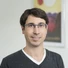Porträt eines Mannes mit Brille, grauer V-Neck-Sweater, heller Hintergrund und einem farbigen Bild im Rahmen dahinter.