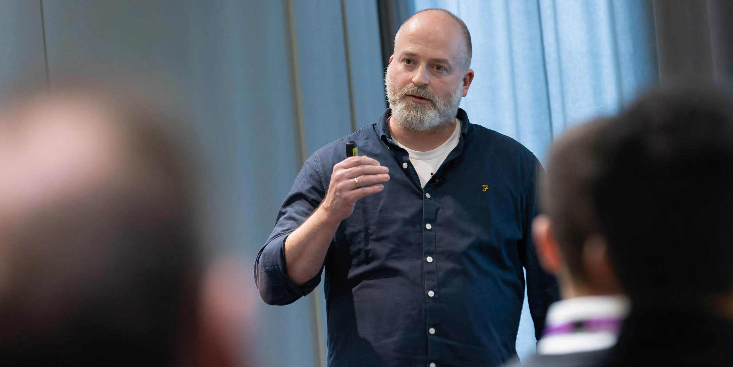 Mann mit dunkelblauem Hemd und grauem Bart spricht vor einer Gruppe; er gestikuliert, im Hintergrund blaue Vorhänge.