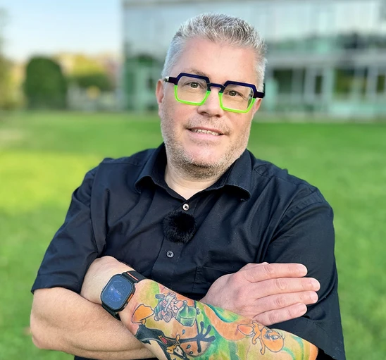 Mann mit grauen Haaren, schwarzem Hemd, grünen Brillenrahmen, farbigem Tattoo am Arm, Arme verschränkt, vor Gras.