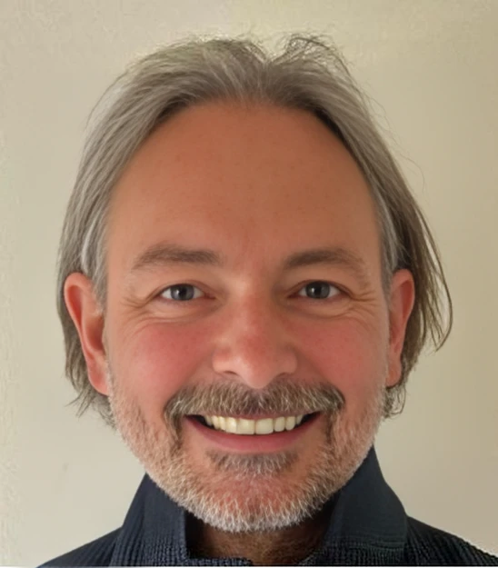 Lächelnder Mann mittleren Alters mit grauem Haar und Bart, von der Schulter aufwärts, vor heller Wand, dunkler Pullover.