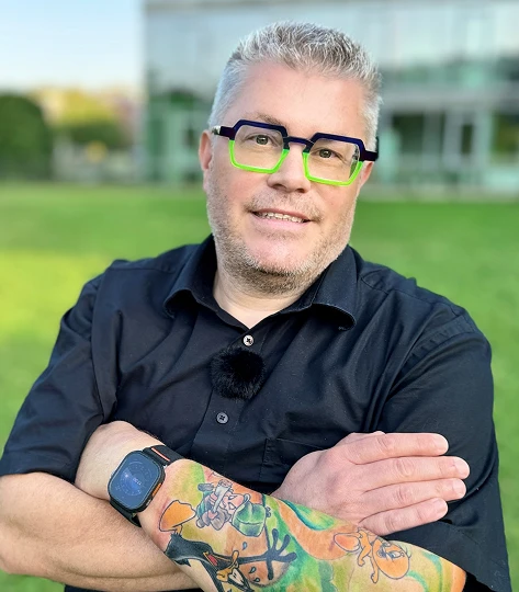 Mann mit grauen Haaren, schwarzem Hemd, grünen Brillenrahmen, farbigem Tattoo am Arm, Arme verschränkt, vor Gras.
