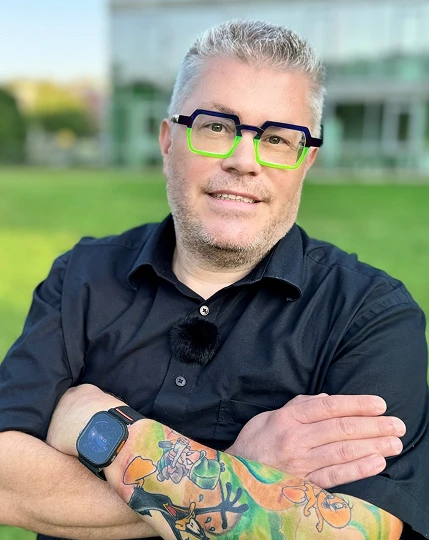 Mann mit grauen Haaren, schwarzem Hemd, grünen Brillenrahmen, farbigem Tattoo am Arm, Arme verschränkt, vor Gras.