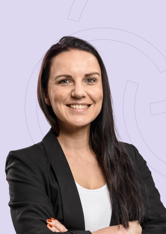 Porträt einer lächelnden Frau mit langen dunklen Haaren, schwarzem Blazer und weißem Oberteil vor hell-lila Hintergrund.