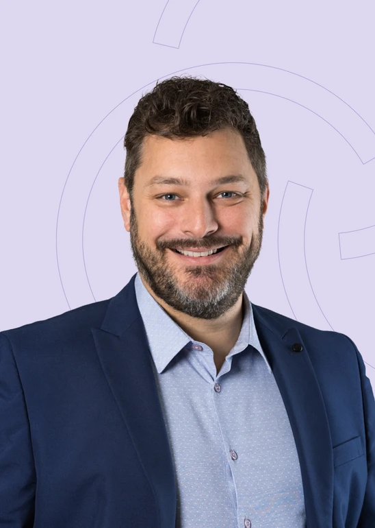 Mann mit dunklem Haar und Bart, blaues Sakko, hellblaues Hemd, vor hellviolettem Hintergrund mit Kreisen.