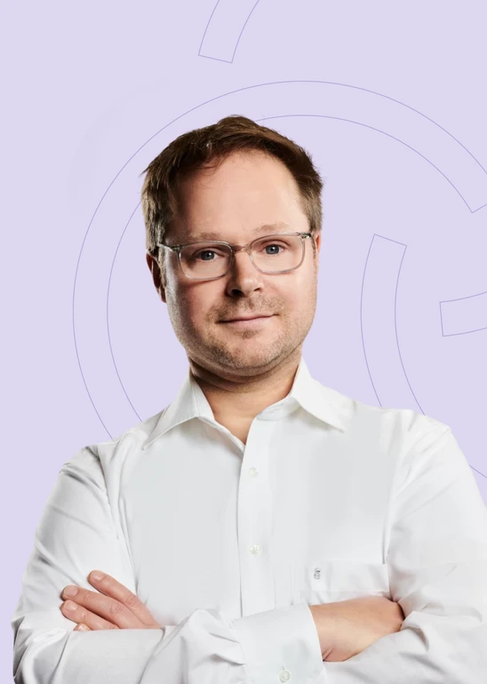 Mann mit Brille, weißes Hemd, verschränkte Arme, vor hellviolettem Hintergrund mit Kreisformen.