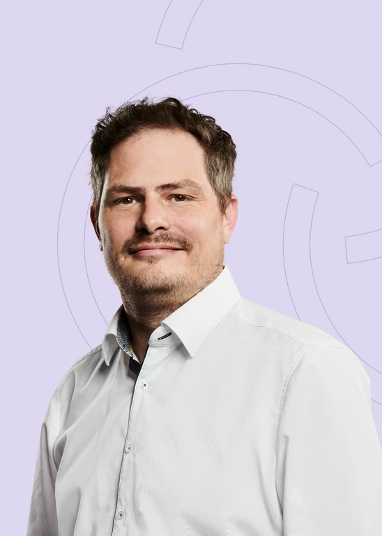 Mann in weißem Hemd vor hellviolettem Hintergrund mit grafischen Kreisen, neutraler Gesichtsausdruck.
